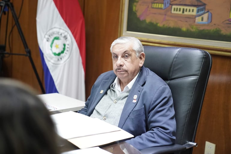 Isaías Ricardo Fretes en chaqueta azul y suéter gris, sentado en oficina con bandera de Paraguay y documentos sobre la mesa.