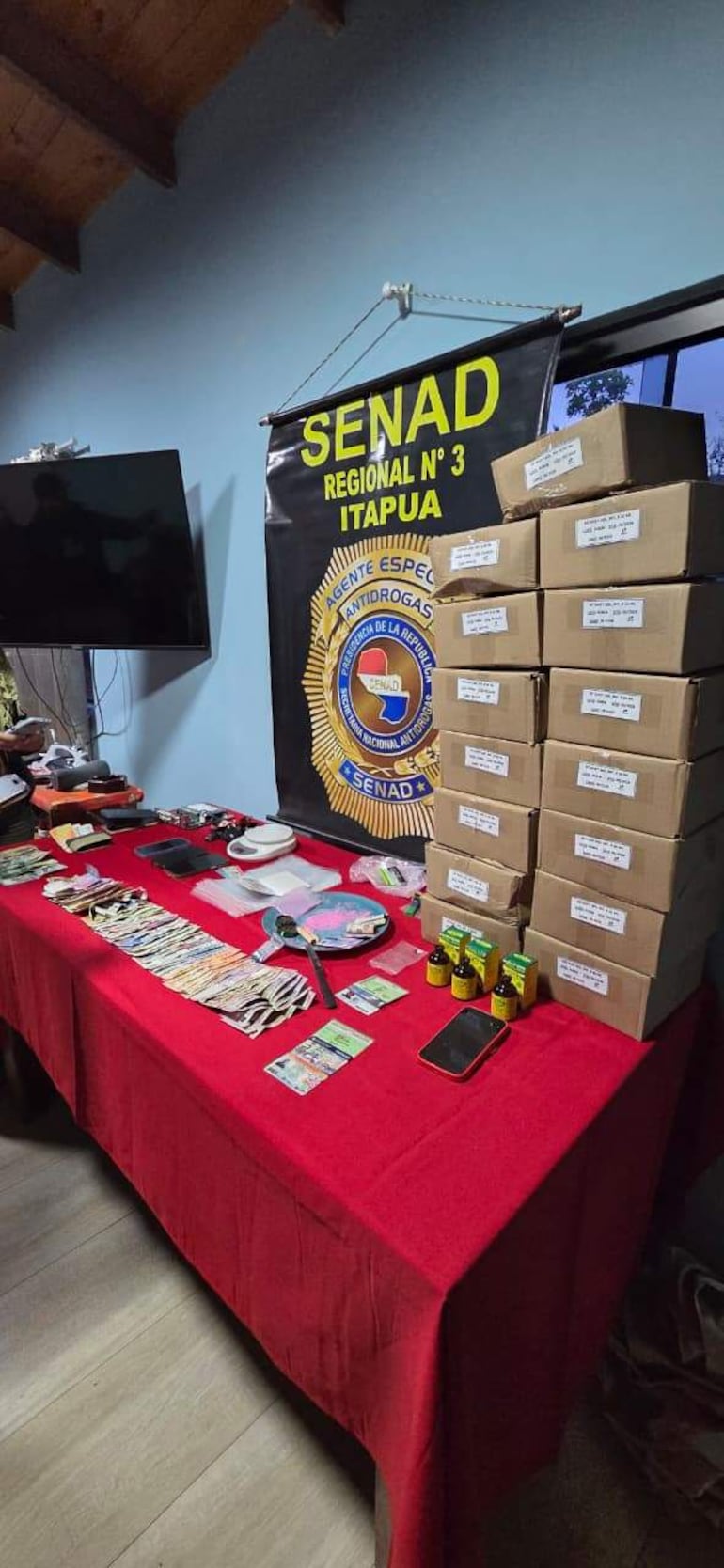 Mesa con billetes, cajas y materiales relacionados con drogas, y banner de la SENAD al fondo.