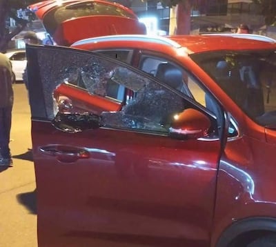Persona de pie junto a un automóvil rojo con la puerta abierta y cristal roto, en un ambiente oscuro con luces de fondo.