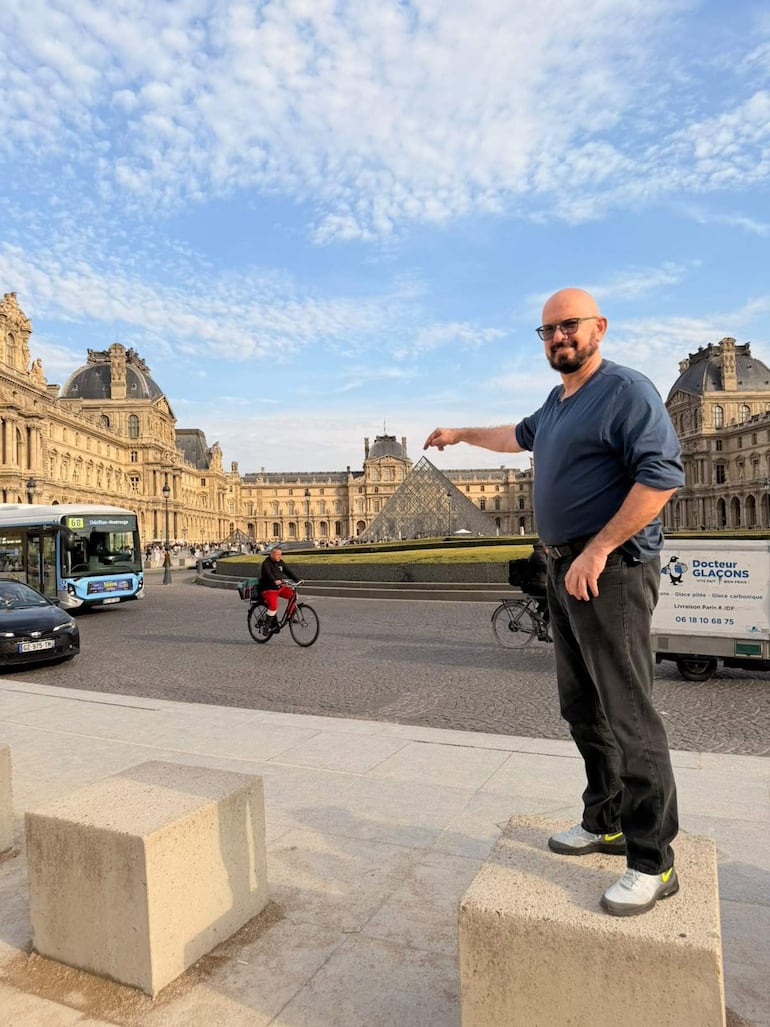 Hombre con camiseta azul y barba señala hacia la pirámide del Louvre, con ciclista y autobús al fondo.