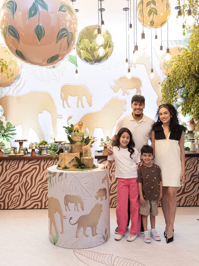 Niña con pantalones rojos y niño en camiseta marrón frente a hombre de camiseta blanca y mujer en vestido negro, todos sonrientes.