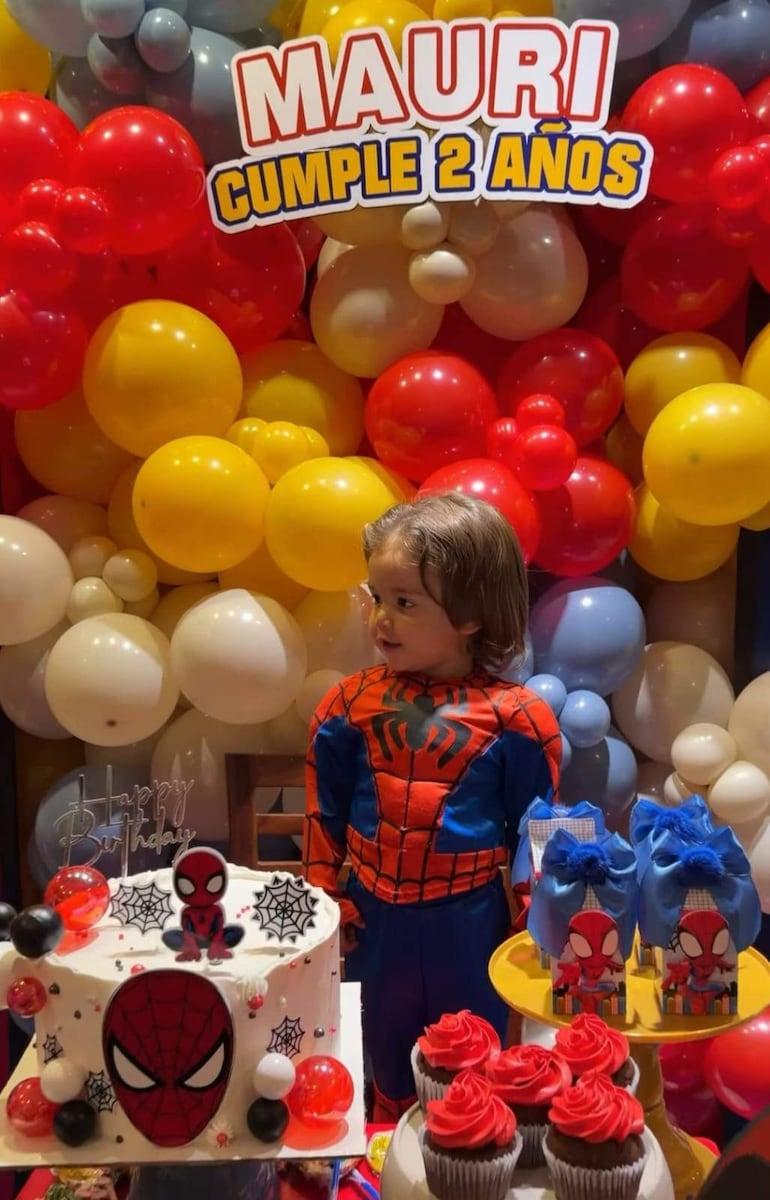 Niño de dos años con traje de Spider-Man sonríe frente a una mesa con pastel y globos coloridos en su fiesta de cumpleaños.