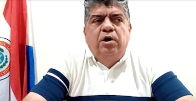 Hombre de mediana edad con cabello oscuro y canoso, en camiseta blanca con franjas azules, hablando frente a una bandera de Paraguay.