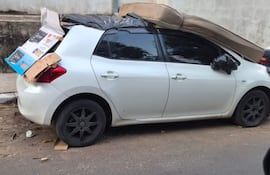 Automóvil blanco estacionado en la calle, con un sofá marrón en el techo y elementos negros adheridos a la parte trasera.