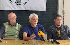 Tres hombres sentados en mesa: uno con gafas y vestimenta verde, otro en camiseta azul con micrófono, y tercero en chaqueta negra.