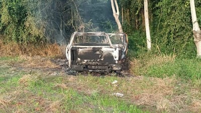 Camioneta quemada en un área rural, rodeada de hierba alta y árboles, con humo saliendo del interior.