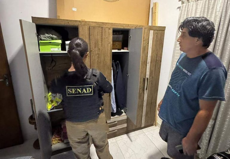 Agentede la SENAD revisa un ropero, mientras un hombre observa en una habitación decorada con colores claros.