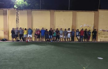 Veinte personas alineadas contra una pared amarilla en un campo deportivo, algunas con brazos cruzados y posturas diversas.