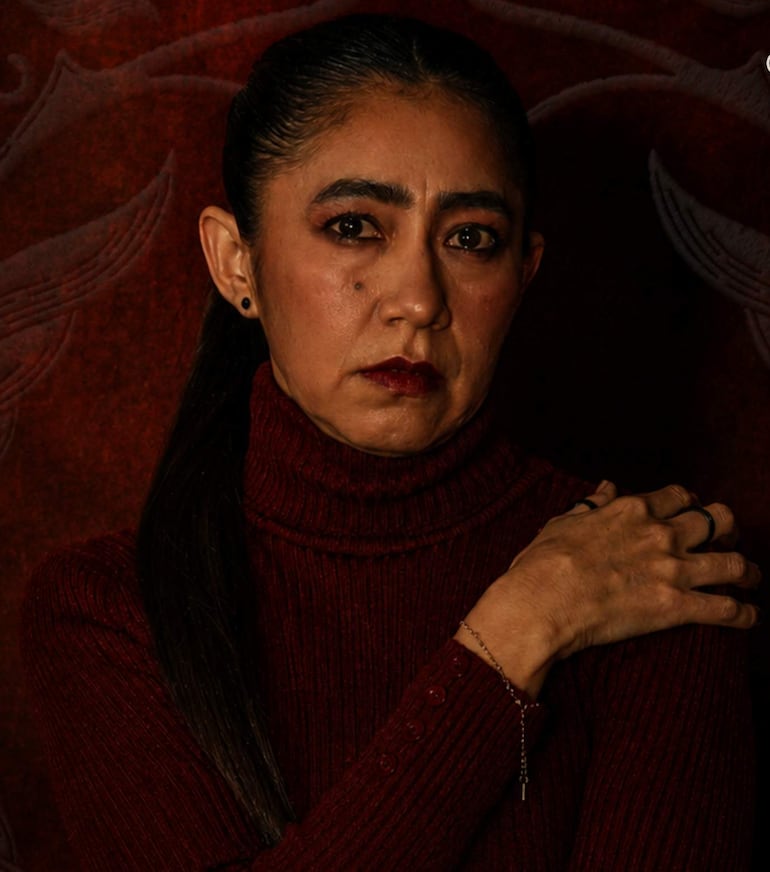 Mujer en suéter rojo oscuro, mirada seria y cabello largo, su mano izquierda en el hombro derecho, fondo oscuro que resalta su figura.