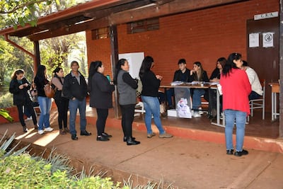 Varias personas esperan en fila en un centro de inscripción; dos mujeres conversan mientras un hombre de chaqueta negra se encuentra al centro.