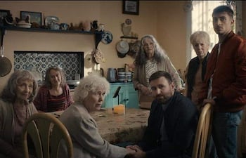 Grupo de siete personas en cocina. Mujer de cabello rizado plateado y hombre sosteniendo la mano de otra mujer, todos con expresiones atentas.