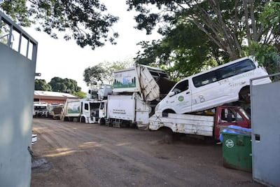 Depósito con camiones de basura apilados, sin personas. Contenedor de basura visible en el entorno sucio.