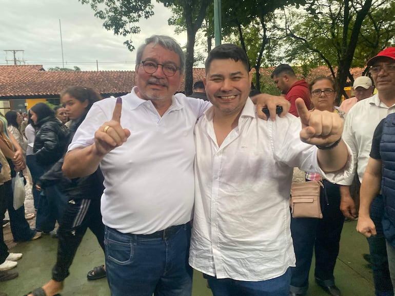 Hombres sonrientes en camisetas blancas levantan un dedo índice, rodeados de un público atento bajo un cielo nublado.