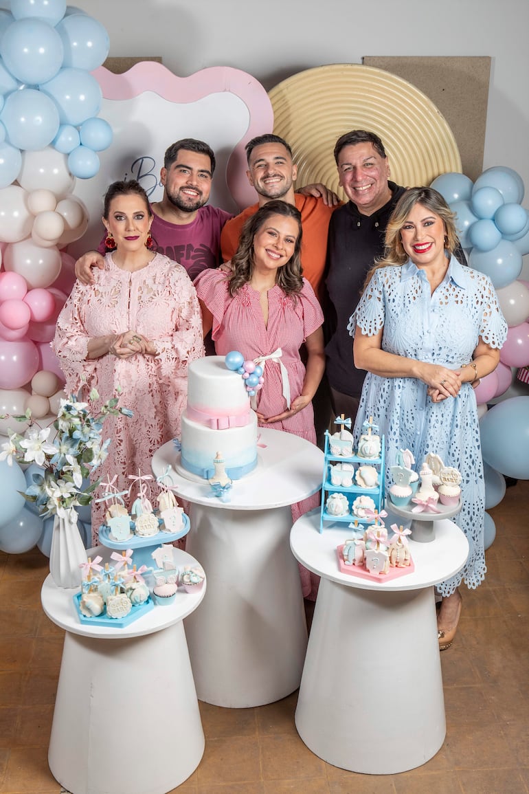 Mujer embarazada al frente, rodeada de amigos sonrientes en un ambiente decorado con globos y mesas de dulces.