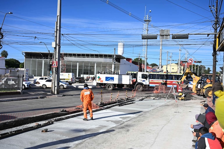 Dos trabajadores en overoles anaranjados, uno en excavadora, rodeados de vehículos en una obra vial al aire libre.