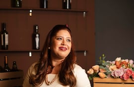 Mujer joven con vestido blanco sonríe en bar, rodeada de botellas y flores de colores suaves.