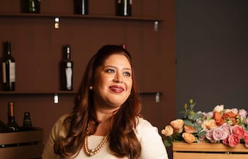 Mujer joven con vestido blanco sonríe en bar, rodeada de botellas y flores de colores suaves.