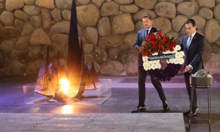 Rubén Ramírez Lezcano y Pedro Alliana con trajes formales, sosteniendo ramo de flores en ceremonia junto a un monumento con llama encendida.