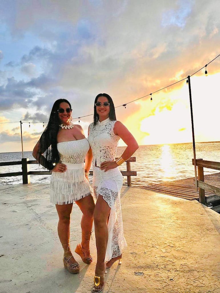 Gloria Vera y Alicia Vera sonríen vestidas de blanco, con gafas de sol, en un entorno cálido y soleado junto al agua.