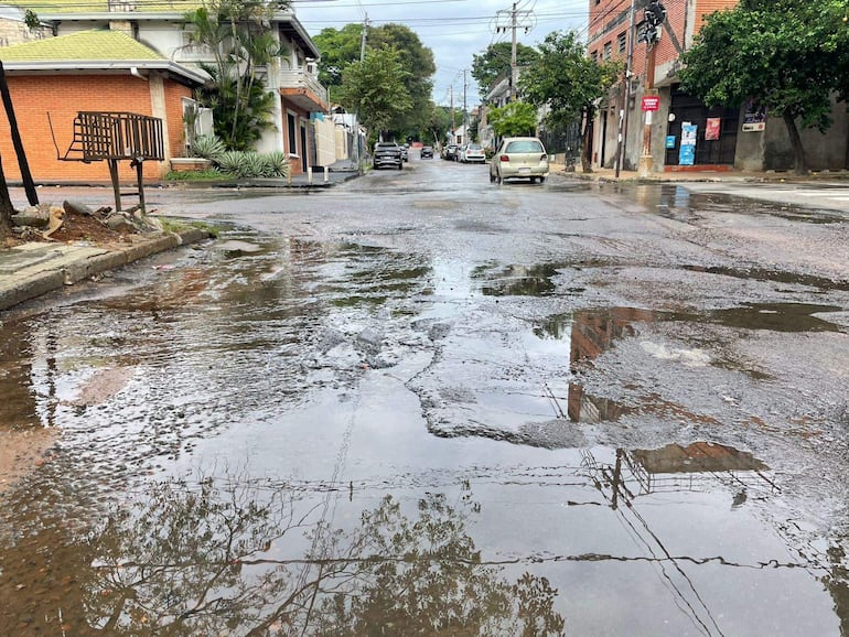 Calle en Tembetary con baches y un charco reflejando árboles y edificios en un día nublado.