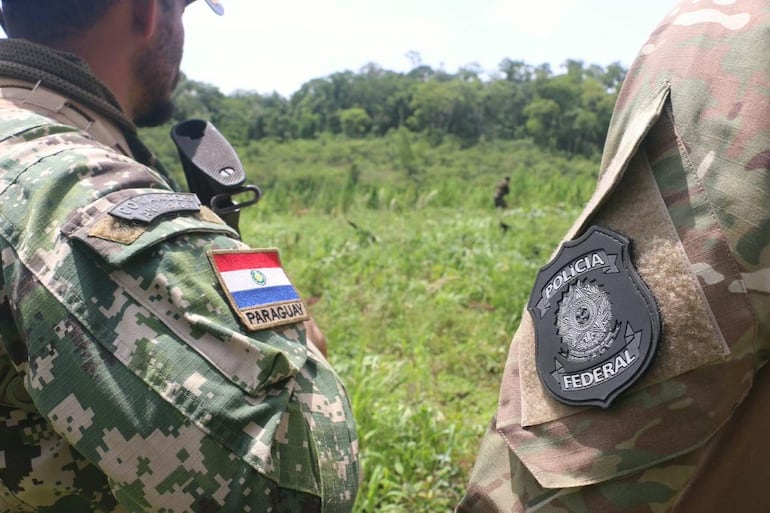 Dos hombres en uniformes de camuflaje, uno con bandera paraguaya, en un entorno natural, sosteniendo armas y en posición de vigilancia.