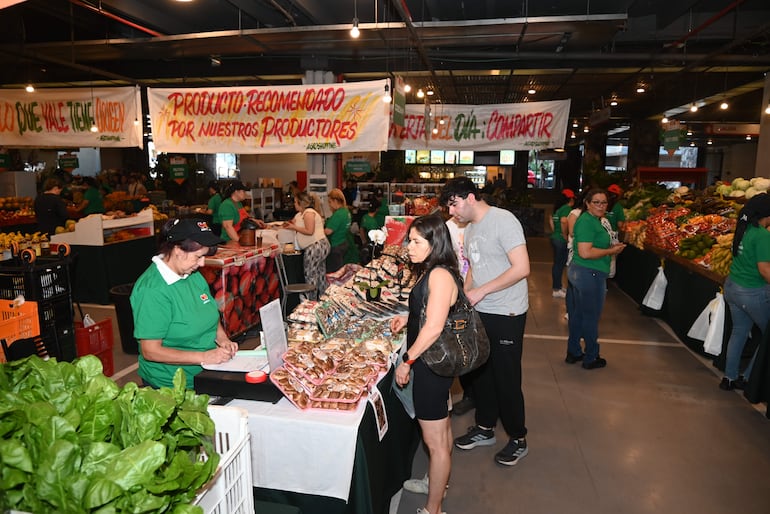 Mujeres y hombres interactúan en un mercado colorido, con productos frescos y un ambiente animado, resaltando la cultura agrícola local.
