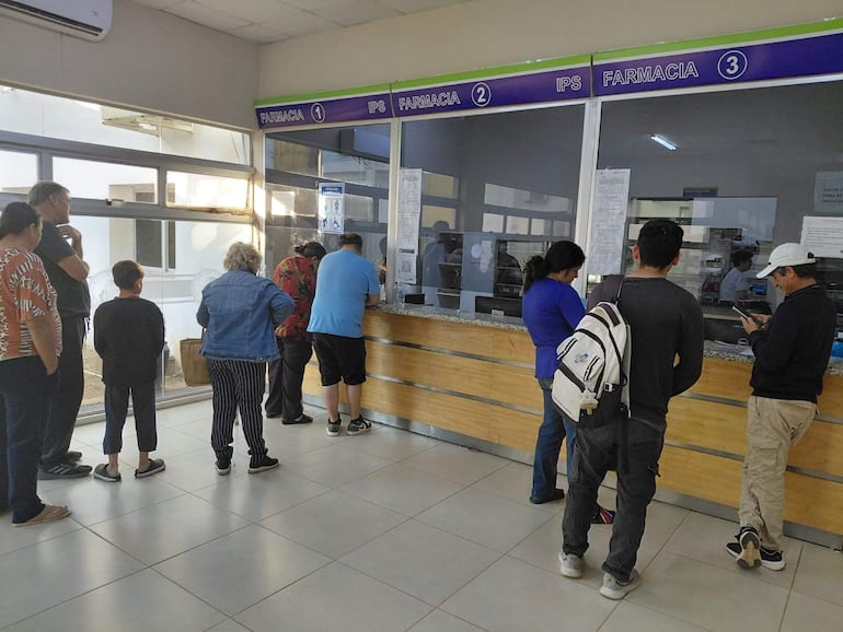Seis personas, una mujer de cabello oscuro y un niño, esperan en fila frente al mostrador de una farmacia IPS iluminada y ordenada.