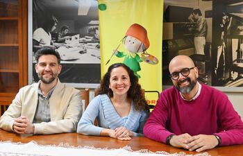 Claudio Pusineri con chaqueta clara, Pili Ortiz en blusa azul y Roberto Galeano Monti en suéter rosado, sonriendo alrededor de una mesa.