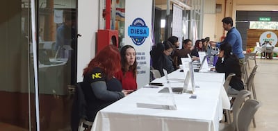 Mujer de cabello largo y rojo conversando en un ambiente interior, con mesas blancas y documentos sobre la mesa.
