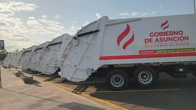 Ocho camiones recolectores de basura en fila, color blanco, con emblema del Gobierno de Asunción en un día soleado.