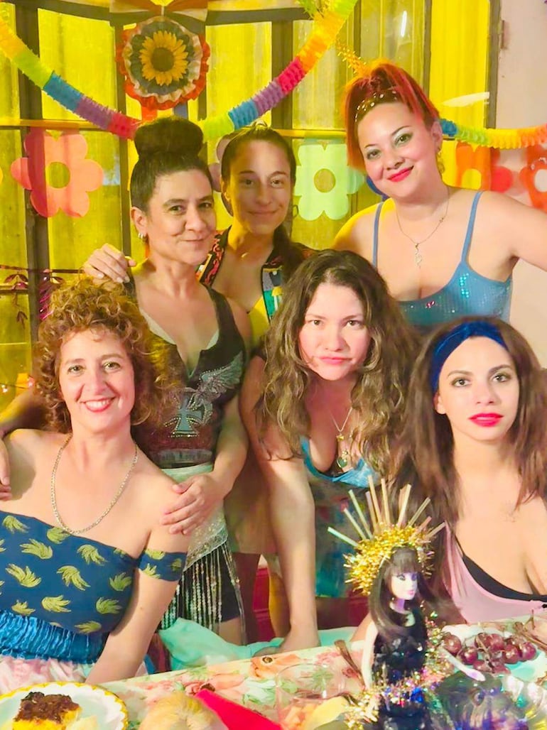 Seis mujeres sonrientes posan en ambiente festivo, con atuendos coloridos y decoración alegre en una mesa.