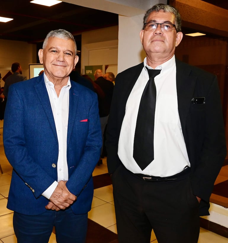 Dos hombres en trajes formales sonríen y conversan, uno con blazer azul y el otro con saco negro y corbata negra, en un salón bien iluminado.