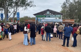 Varios adultos y niños en fila, vestidos con abrigos, conversan mientras esperan, rodeados de árboles y carteles de boletería.