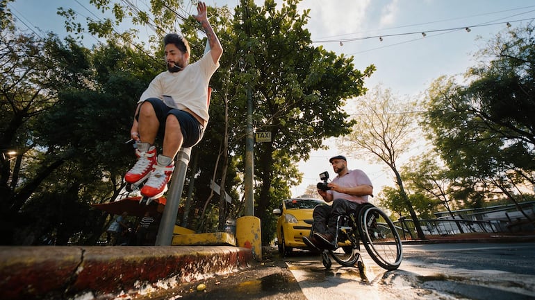 Hombre con camiseta clara y patines rojos hace piruetas, mientras un espectador en silla de ruedas lo graba, fondo urbano.