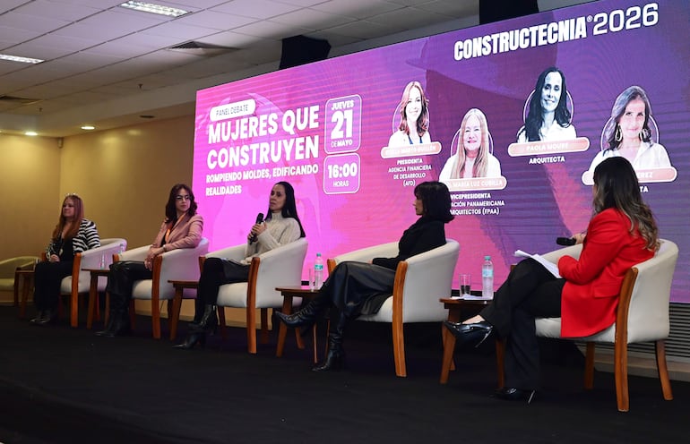 Cinco mujeres en sillas blancas, una habla mientras las demás escuchan atentamente, en un auditorio decorado con un fondo colorido.