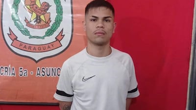 Hombre joven con camiseta blanca de Nike y expresión seria, frente a un escudo institucional rojo de Paraguay.