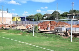 La imagen muestra un campo de fútbol en primer plano, con una portería a la izquierda y césped bien cuidado. En el fondo, hay un sitio de construcción donde se observan varias personas trabajando. Aproximadamente hay seis a ocho trabajadores, algunos de los cuales están en posiciones de pie, realizando tareas de excavación y transporte de materiales.
No hay figuras públicas reconocibles en la imagen. En cuanto a elementos visuales que identifiquen un equipo de fútbol, no se observan camisetas, escudos o colores institucionales.
El entorno es exterior y parece ser un área cercana a un complejo deportivo, dado que hay edificios residenciales y comerciales visibles en el fondo. Las acciones en curso incluyen la construcción y el movimiento de materiales como arena y ladrillos. No hay texto visible en la imagen.