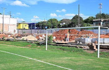 La imagen muestra un campo de fútbol en primer plano, con una portería a la izquierda y césped bien cuidado. En el fondo, hay un sitio de construcción donde se observan varias personas trabajando. Aproximadamente hay seis a ocho trabajadores, algunos de los cuales están en posiciones de pie, realizando tareas de excavación y transporte de materiales.
No hay figuras públicas reconocibles en la imagen. En cuanto a elementos visuales que identifiquen un equipo de fútbol, no se observan camisetas, escudos o colores institucionales.
El entorno es exterior y parece ser un área cercana a un complejo deportivo, dado que hay edificios residenciales y comerciales visibles en el fondo. Las acciones en curso incluyen la construcción y el movimiento de materiales como arena y ladrillos. No hay texto visible en la imagen.