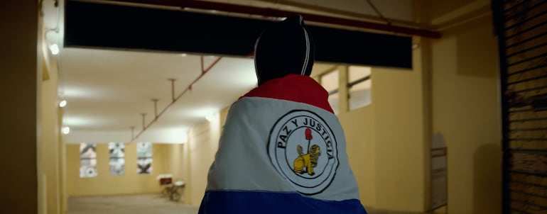 Persona de espaldas envuelta en bandera paraguaya, con logo de KIA en el pasillo iluminado del edificio.
