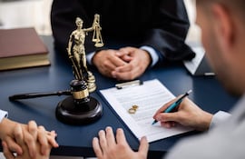 Persona con vestimenta formal escucha mientras otra firma documentos sobre una mesa con martillo de juez y estatua de la Justicia.