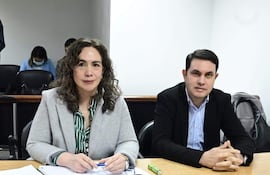 Alba Zaracho con cabello rizado y abrigo gris escribe mientras Hernán Rivas en camisa de cuadros escucha atentamente en ambiente formal.