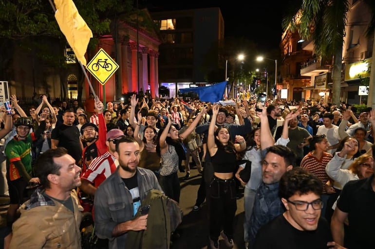 Multitud vibrante durante una celebración, personas levantando los brazos y banderas, iluminación festiva en el fondo.