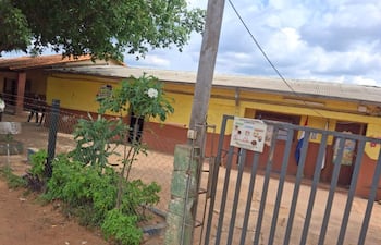 Escuela pública de un piso con paredes amarillas y jardín, cerca de metal visible pero sin personas presentes.