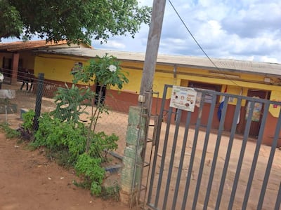 Escuela pública de un piso con paredes amarillas y jardín, cerca de metal visible pero sin personas presentes.