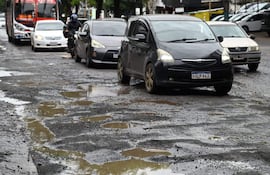 Calle con baches y charcos, coches y un autobús en movimiento, entorno urbano con árboles y comercios al fondo.