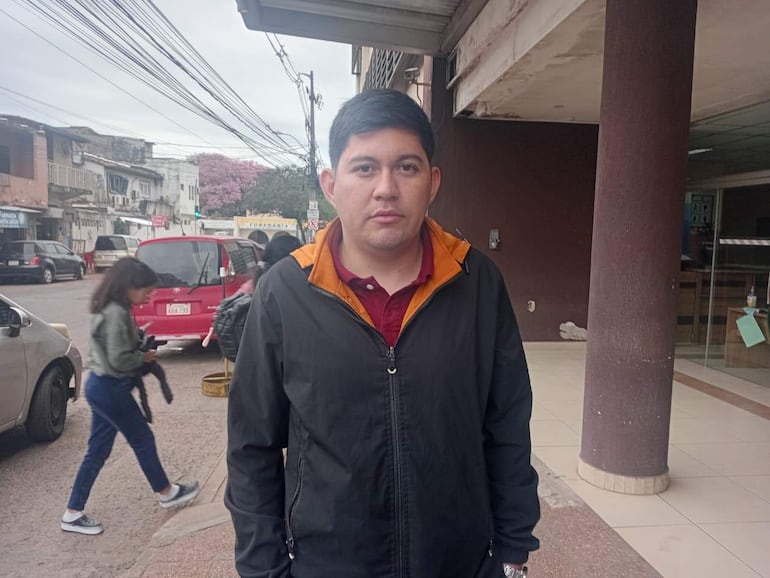 Hombre de pie con chaqueta negra y mujer con chaqueta verde caminando en una calle urbana, rodeados de vehículos y edificios.