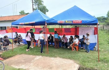 Grupo de personas bajo carpas azules discutiendo, con expresión de seriedad, rodeados de carteles y una bandera paraguaya.