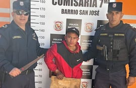 Tres hombres en la comisaría: dos policías con gafas de sol, uno sostiene un machete, el detenido viste camiseta roja y negra, rostro tranquilo.