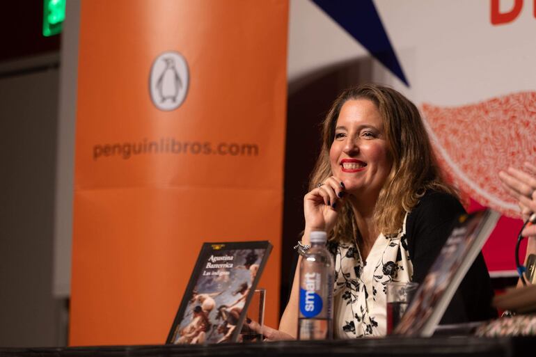 Agustina Bazterrica sonríe sentada frente a una mesa con sus libros y un vaso de agua en un evento literario.