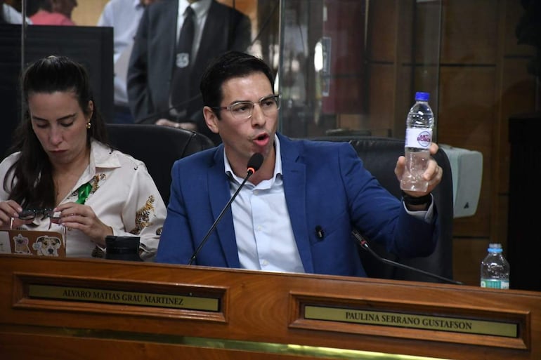 Alvaro Grau en traje azul sosteniendo una botella de agua, acompañado de Jazmin Galeano revisando documentos en un ambiente formal.